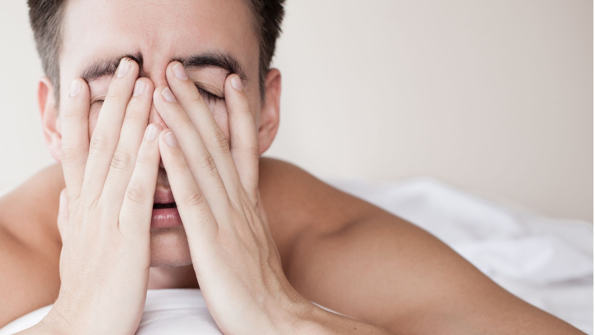 A man with his eyes closed lies on his bed on his front, propped up by his arms, and covers his face with his hands.