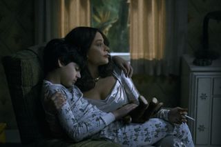 a woman in a silver dress (Sofia Vergara as Griselda) sits in a chair and holds her young son (Martin Fajardo as Ozzy), who is wearing white printed pajamas