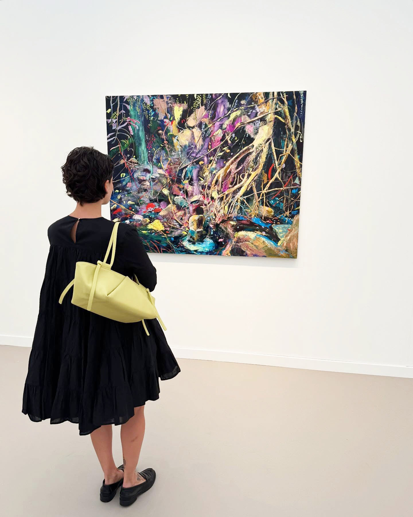 A woman wearing a black dress, yellow hand, and black flats in front of a painting