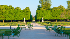a garden with large green trees and dirt walkways with green metal chairs on either side 