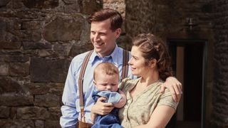 Nicholas Ralph in a blue shirt as James stands with Rachel Shenton in a beige blouse holding baby Jimmy in All Creatures Great and Small.