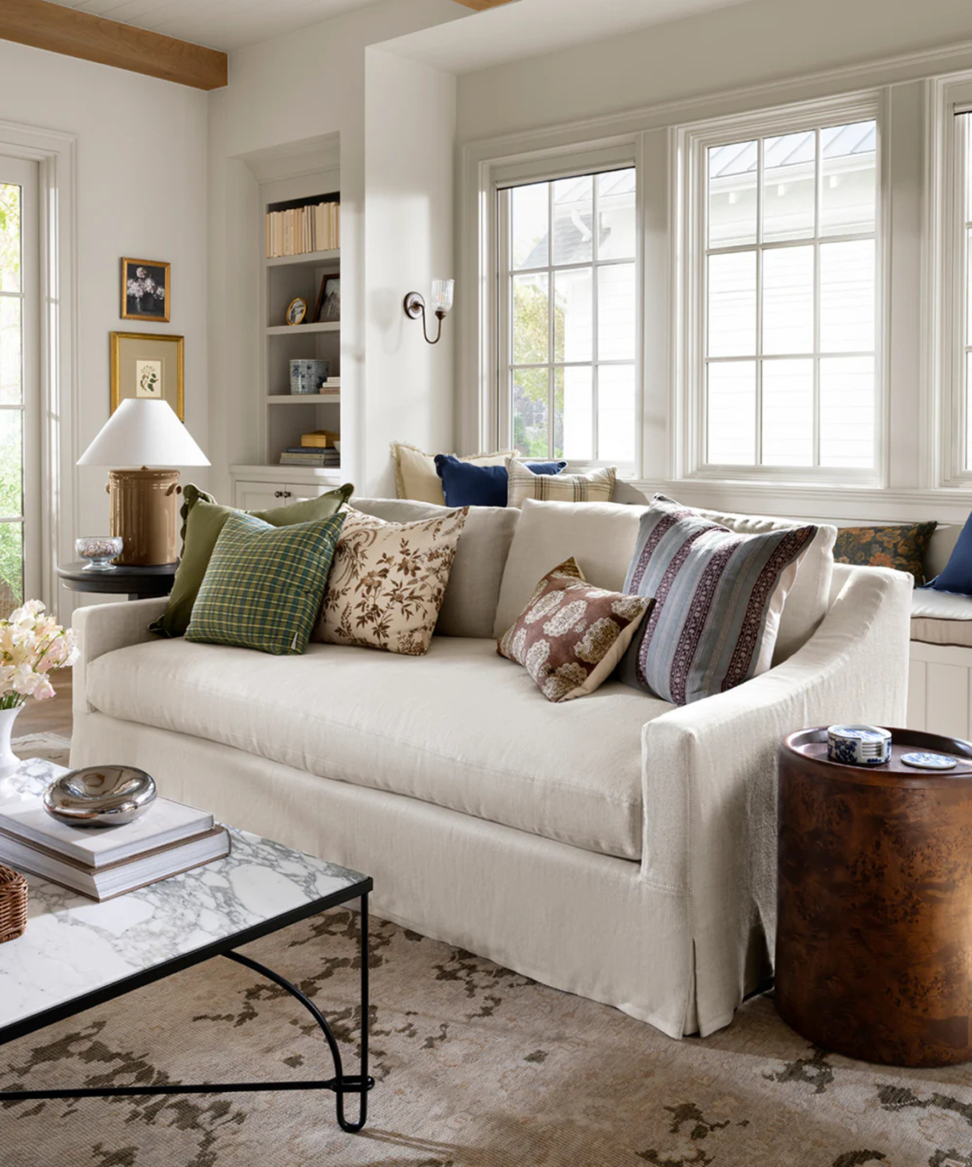 a living room with a couch with several pillows on top, a burl wood side table, and a coffee table with a chrome trinket dish on top