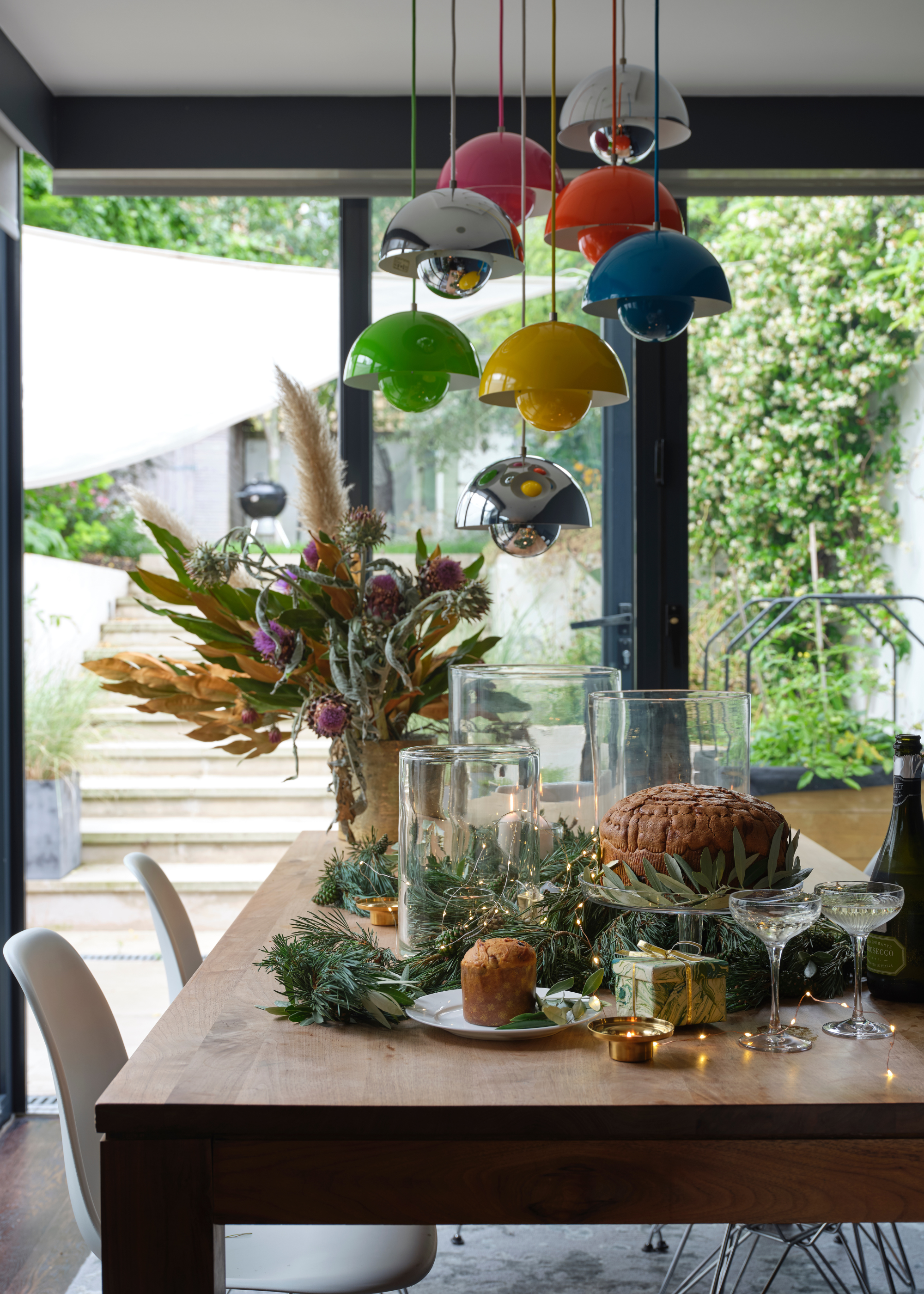A Christmas table with hurricane lanterns surrounded by foliage and string lights with a Christmas cake on display and colorful pendant lighting hanging above with sliding doors leading to the garden in the background