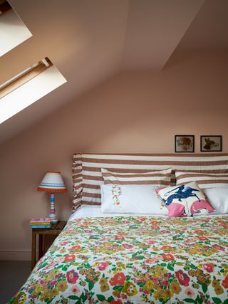 Ένα ροζ υπνοδωμάτιο στη σοφίτα με θολωτή οροφή και δύο φεγγίτες που στεγάζουν ένα κρεβάτι με ριγέ κεφαλάρι και κλινοσκεπάσματα με λουλούδια
