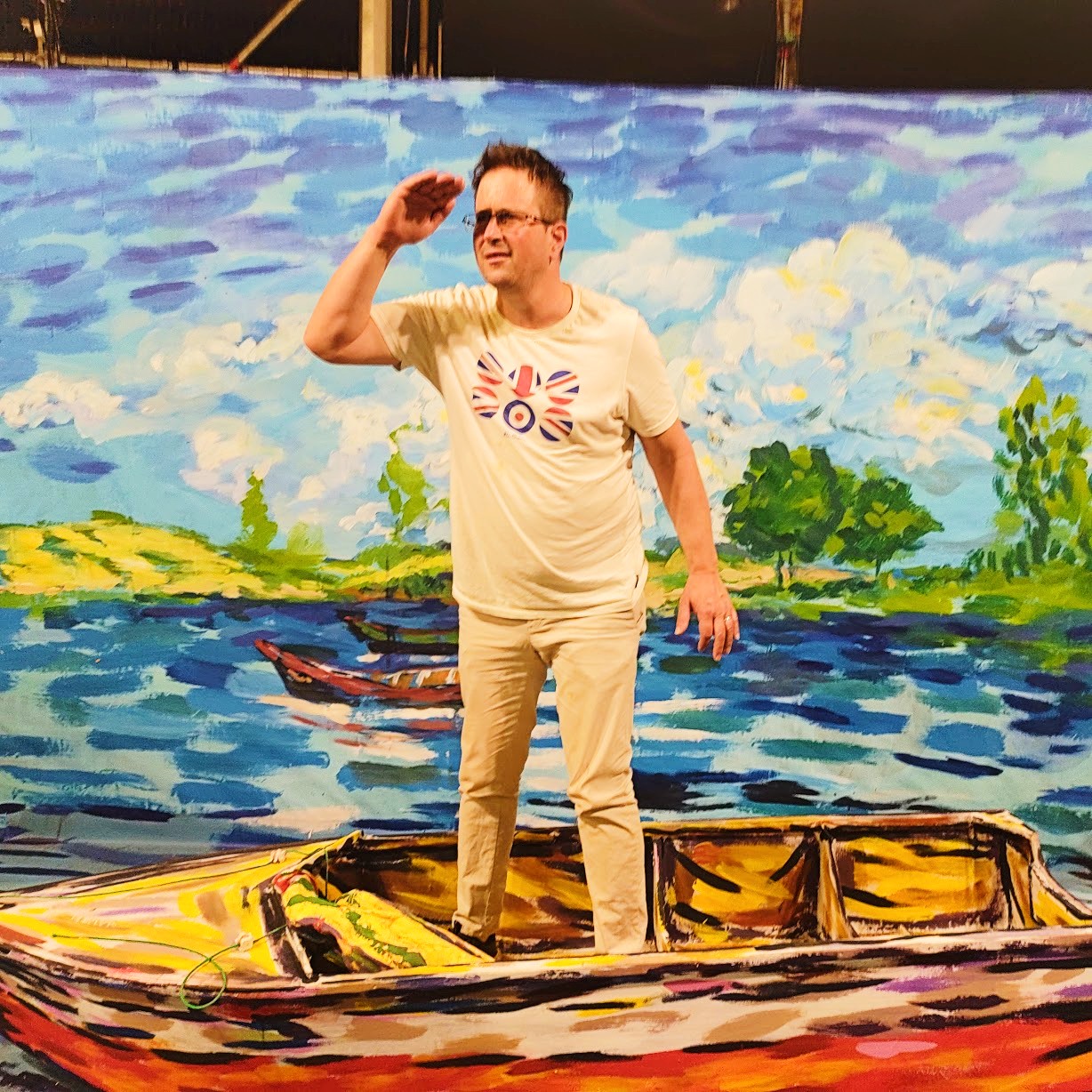 A man in light-colored clothing and a graphic tee stands inside a painted boat, posing with his hand to his forehead against a vibrant, impressionistic lake backdrop.