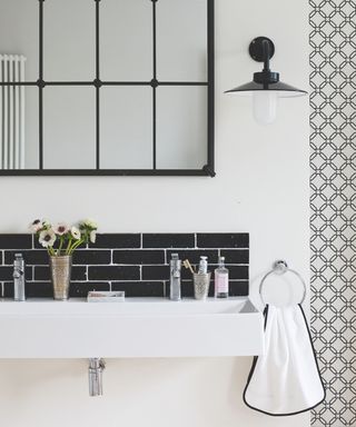 A black and white bathroom with a large white sink with black tiled backsplash, flowers and cosmetics with silver faucets, a black industrial style mirror and light fixture, a white towel with black trim on a hand towel holder and geometric black and white patterned wallpaper to the right