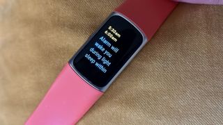 The Fitbit Charge 6 sitting on a pillow, showing an alarm window of 5:30-6:00am, then the words "Alarm will wake you during light sleep within..." (the words cut off)