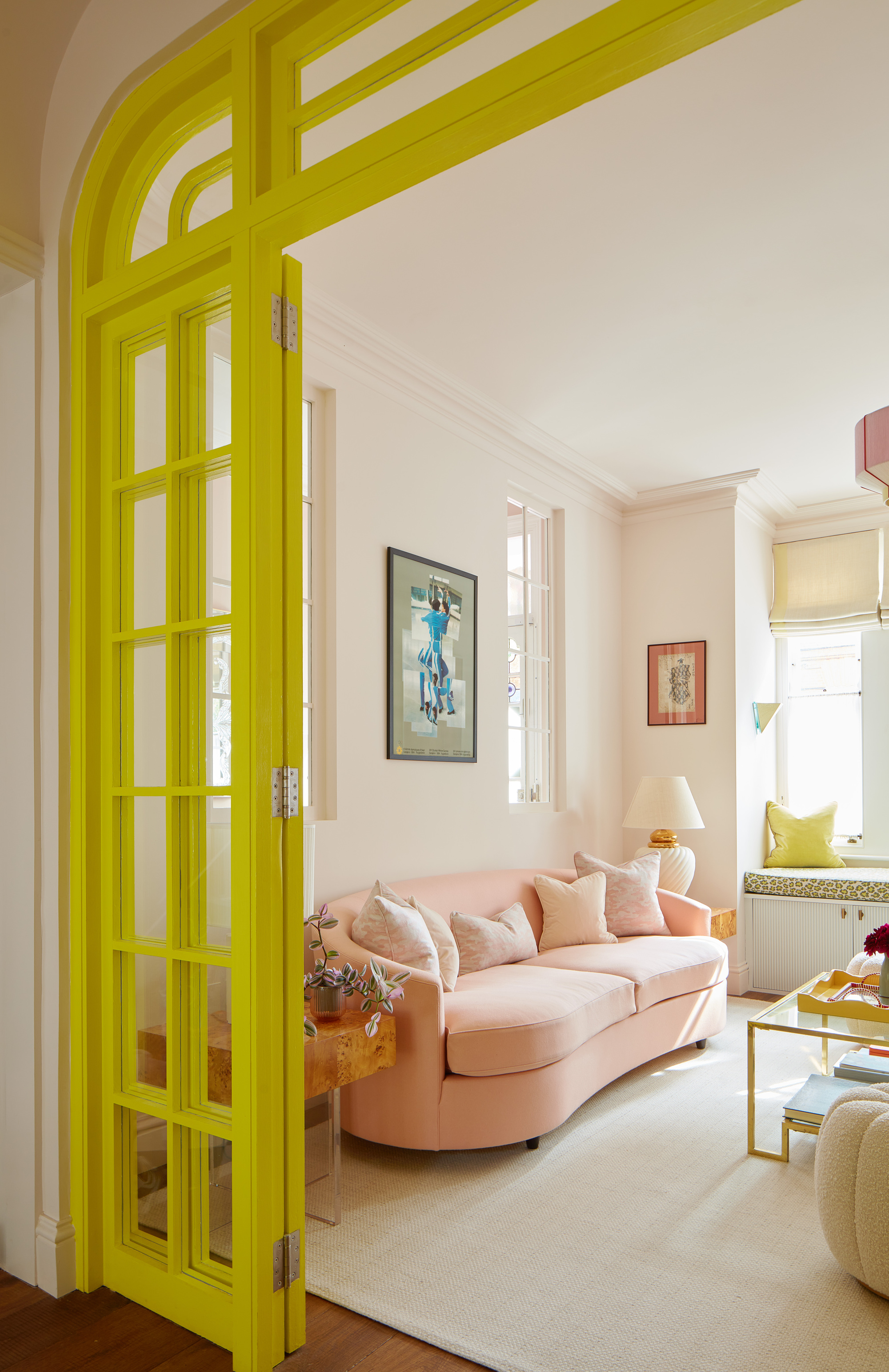 muted pastel living room with pink curved sofa, white walls, white floor rug, artwork on walls, and door frame painted in neon yellow to contrast