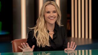 A press image of Reese Witherspoon sitting behind a desk smiling with her hands slightly lifted off the table in Season 3 of The Morning Show.