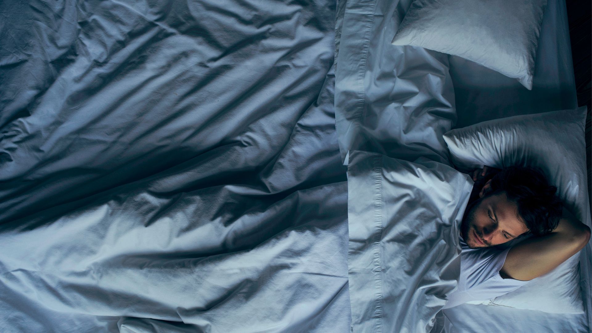 A man lies awake at night. He is lying on his back in a bed with white bedding.