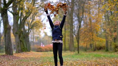 Healthy older woman jumps up in air creating a heart shape with fallen leaves