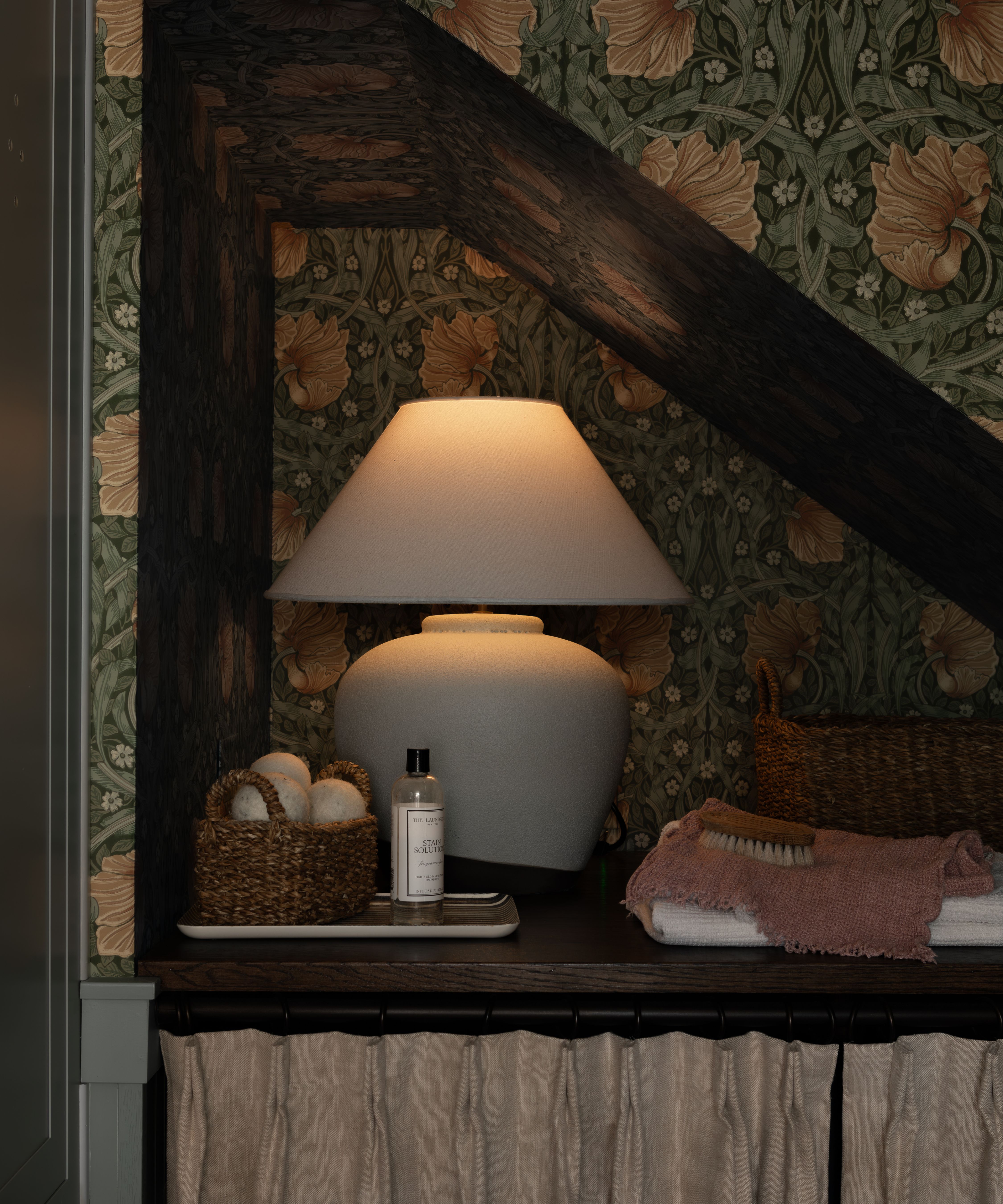 a wallpapered angled storage nook in a small bathroom with a lamp and basket storage