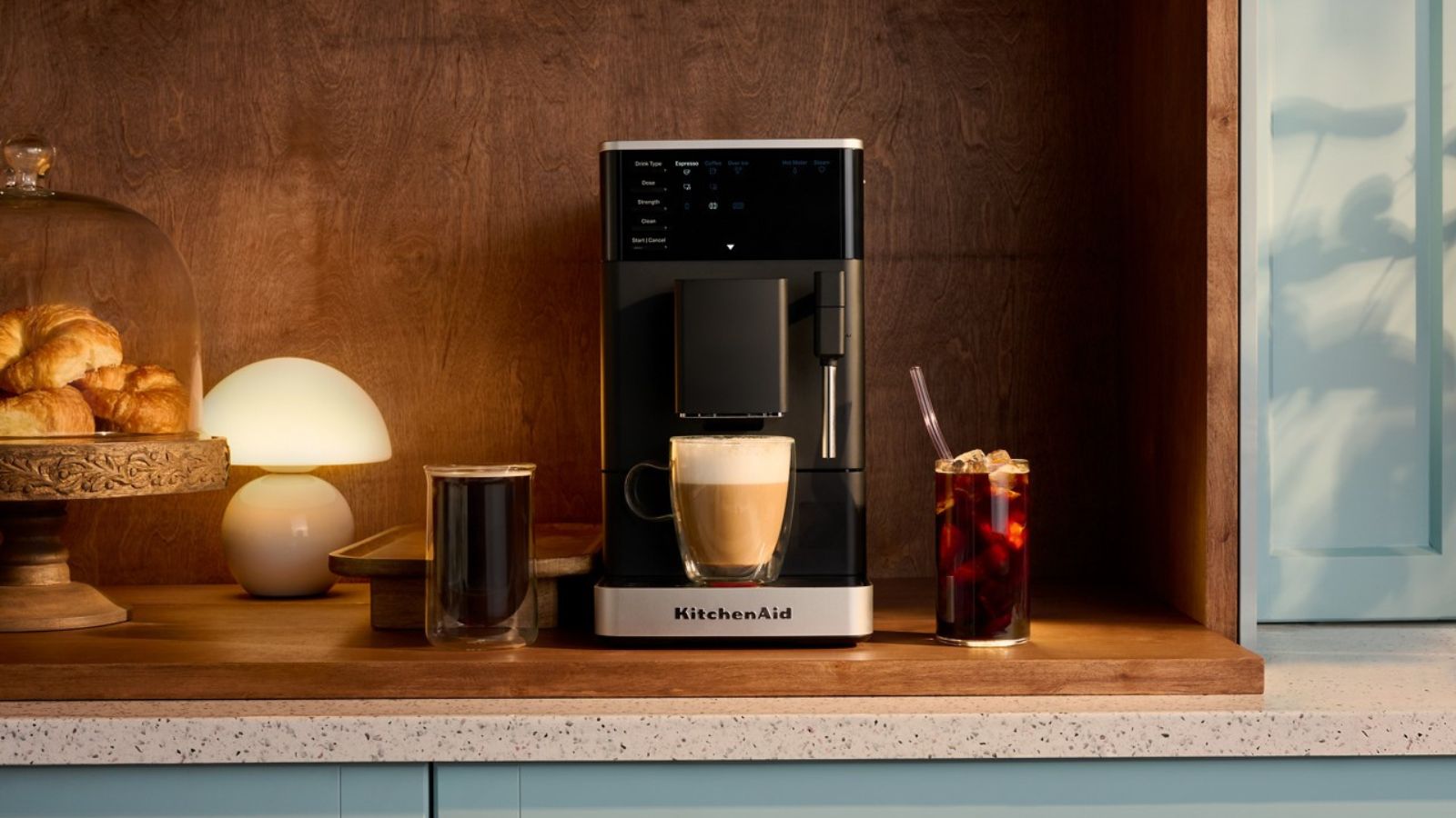 A black compact KitchenAid coffee maker on a wooden counter with a wooden backsplash, with a lamp and a cake stand full of pastries beside it. 