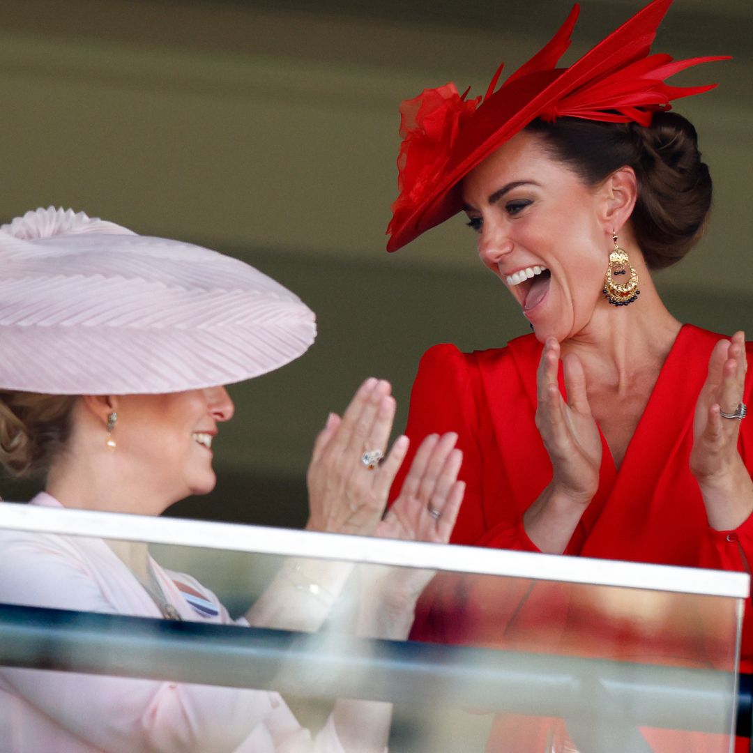 Kate Middleton wearing a red hat and dress laughing and clapping while looking at Duchess Sophie, wearing a pink dress and hat, at Royal Ascot