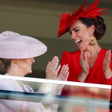 Kate Middleton wearing a red hat and dress laughing and clapping while looking at Duchess Sophie, wearing a pink dress and hat, at Royal Ascot