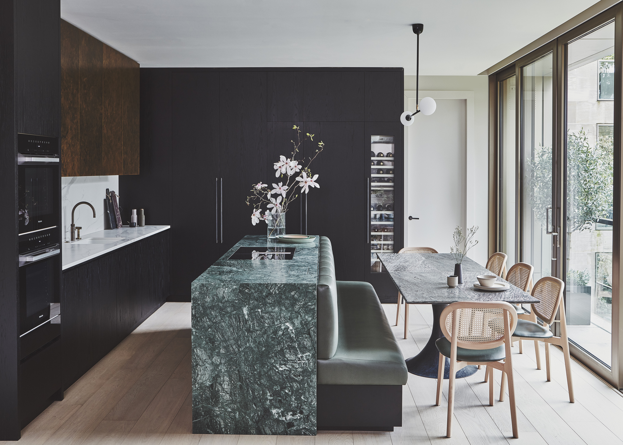 A kitchen with black cabinetry, an island with built-in banquette seating, a dining table, and a wine fridge with a glass door built-into the cabinets
