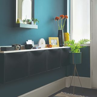 A blue-painted hallway with black IKEA shoe cabinets and a floating shelf on top displaying decorative items and a mirror with a shelf hung above