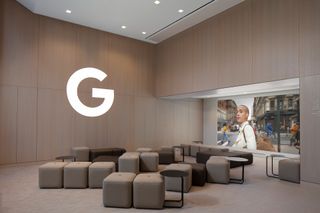 Google New York Store's workshop space, with wooden walls and upholstered seats in beige and brown, arranged in front of a large screen. On the wall is an oversized and illuminated "G" from the Google logo