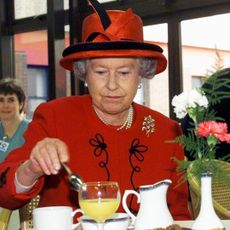 Queen Elizabeth in a red coat and hat stirring tea at a table