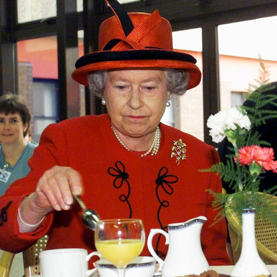 Queen Elizabeth in a red coat and hat stirring tea at a table