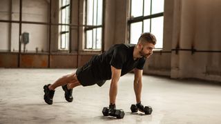 Man in black activewear in a warehouse performing a plank hold while holding two light dumbbells during dumbbell workout