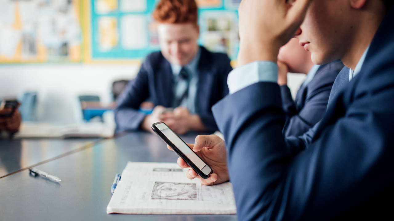 A child sitting in a classroom scrolls on his phone at his desk