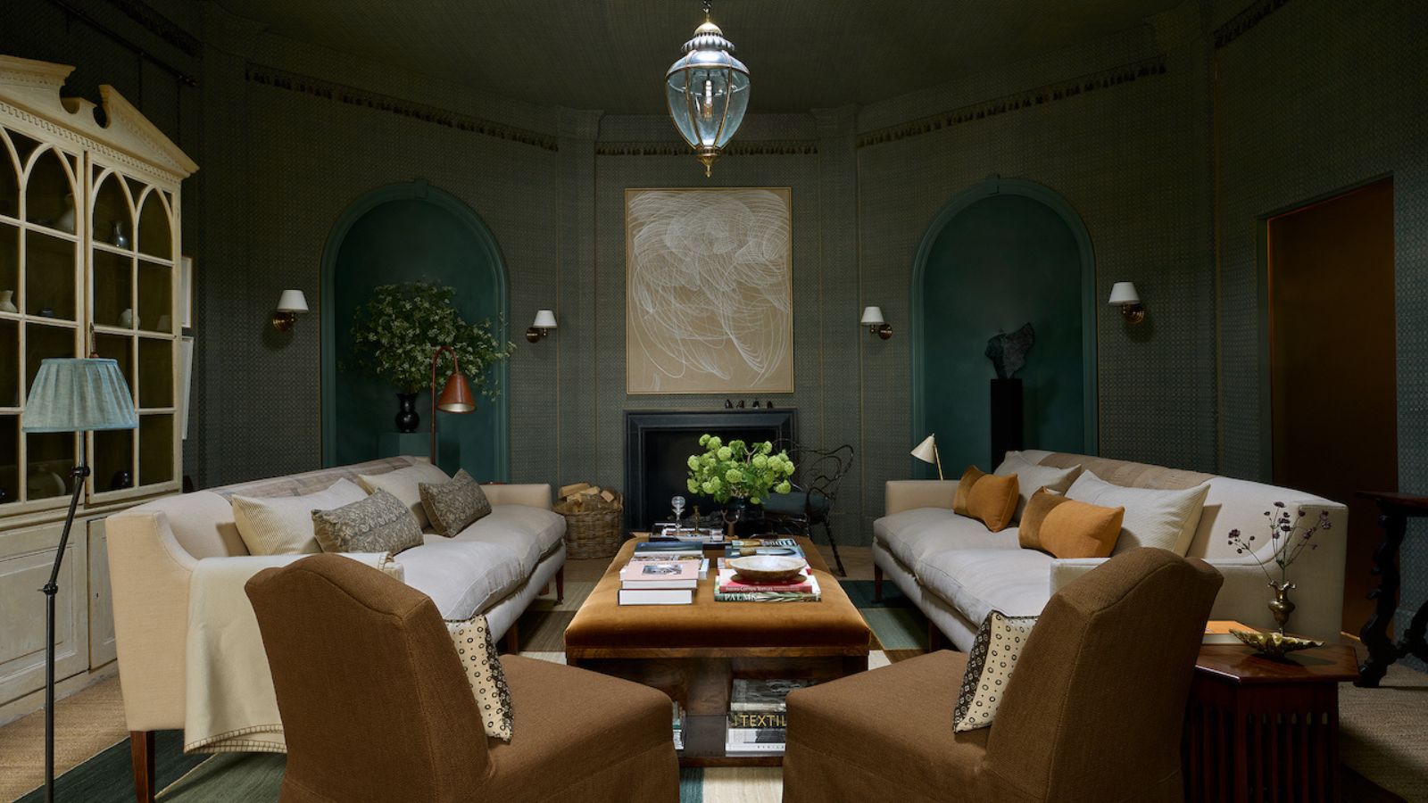 Living room with blue grasscloth walls, alcoves with statues, a velvet brown coffee table with books on top, cream linen sofas, slipcovered brown armchairs, am armoire with books and a glass pendant