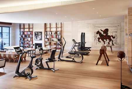 A gym area with various exercise equipment on a wooden floor with a sitting area in front of it with bookshelves and a fireplace on the wall.