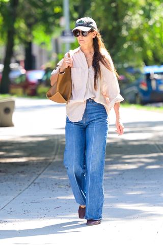 Katie Holmes wearing a baseball cap, oversized button-down shirt, brown tote bag, wide-leg jeans, and burgundy flats.