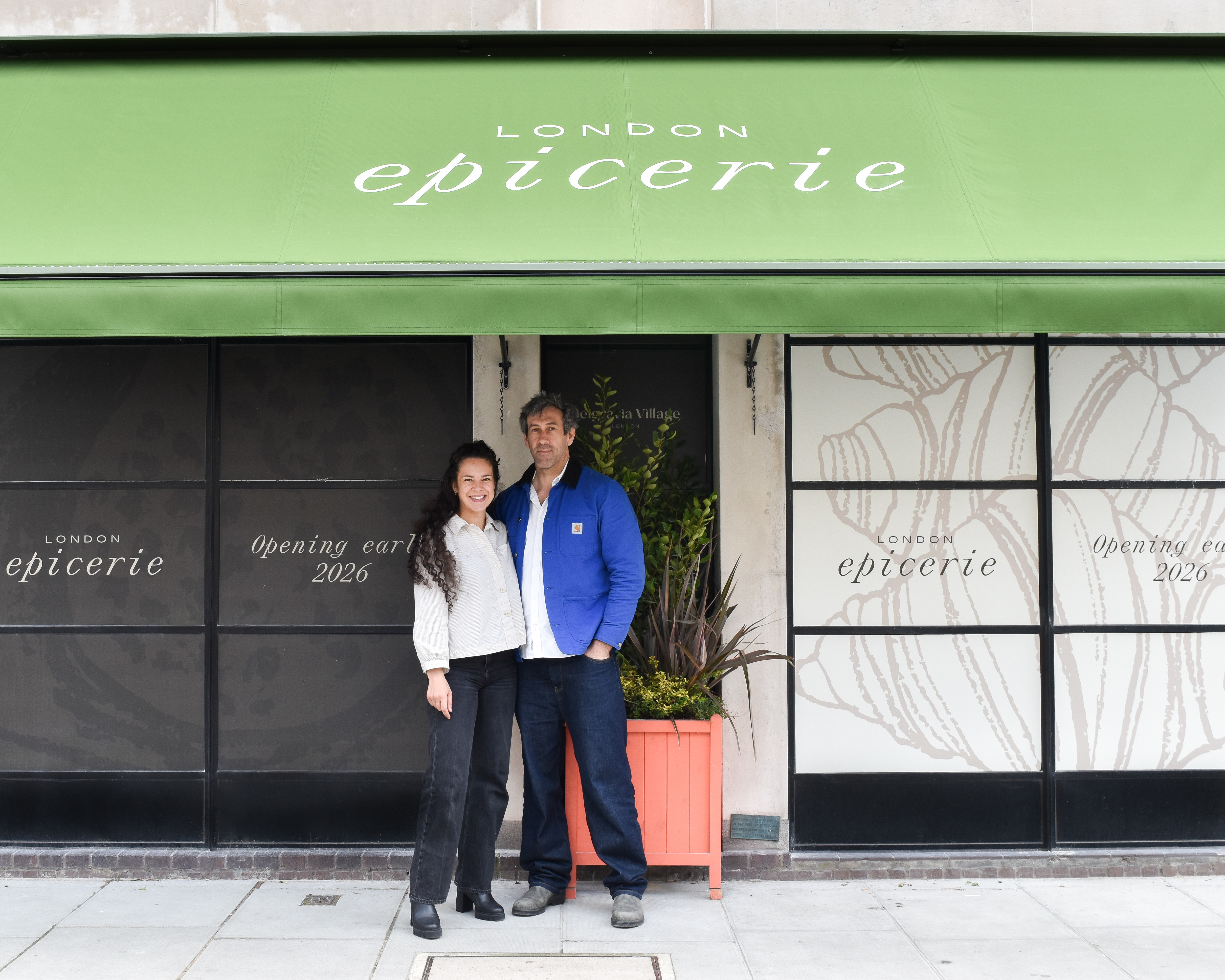 A young couple wearing dark blue trousers, and a gray and a blue jacket, respectively, stands before a yet-to-be-opened "London Epicerie" shop front under a green awning.