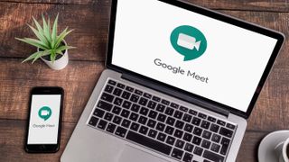 Google Meet logo being displayed on a laptop and a phone that are resting on a wooden table