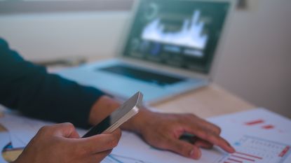 A man does financial planning with paperwork, his phone and a laptop, only his hands showing.