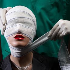 a woman in red lipstick with a head wrap and a surgeon behind her wrapping her face