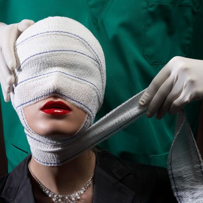 a woman in red lipstick with a head wrap and a surgeon behind her wrapping her face