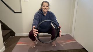 Jennifer Rizzuto performs a squat and chest press with a Pilates ring. She is squatting down with knees bent, arms extended in front of her, holding a Pilates ring.