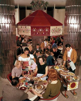 Busy restaurant image in 1960, customers sat eating at tables, red chairs, waitress and waiters serving food, food trolley, neutral floor, tall metallic pillars, red decorated canopy above a table