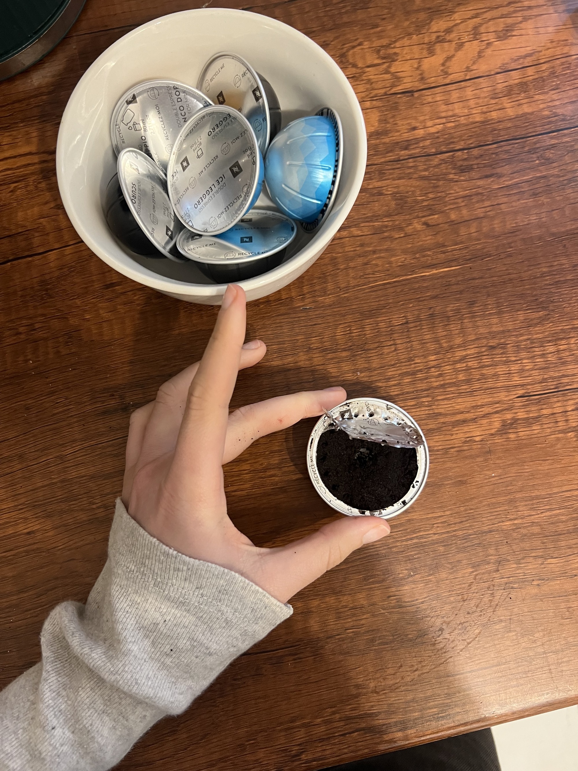 image of a hand wearing a long-sleeved gray shirt holding an open coffee pod over a wooden countertop. There is a bowl of coffee pods also on the counter.