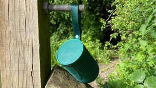 En grön Bang & Olufsen Explore-högtalare hänger i sin tillhörande rem på ett trästaket utomhus, med en somrig grön bakgrund.