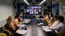 A board meeting in a modern office, with everyone in the room looking at a screen showing remote working colleagues.