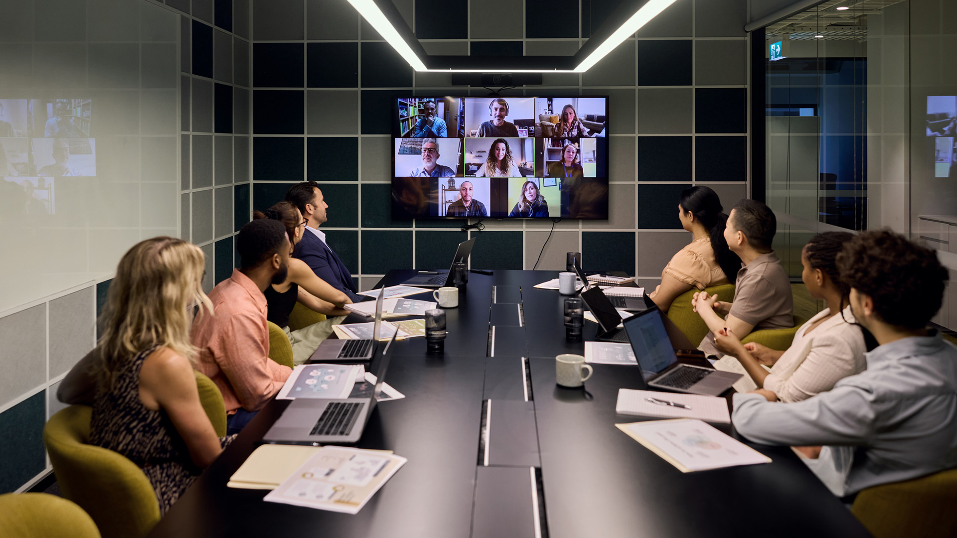A board meeting in a modern office, with everyone in the room looking at a screen showing remote working colleagues.