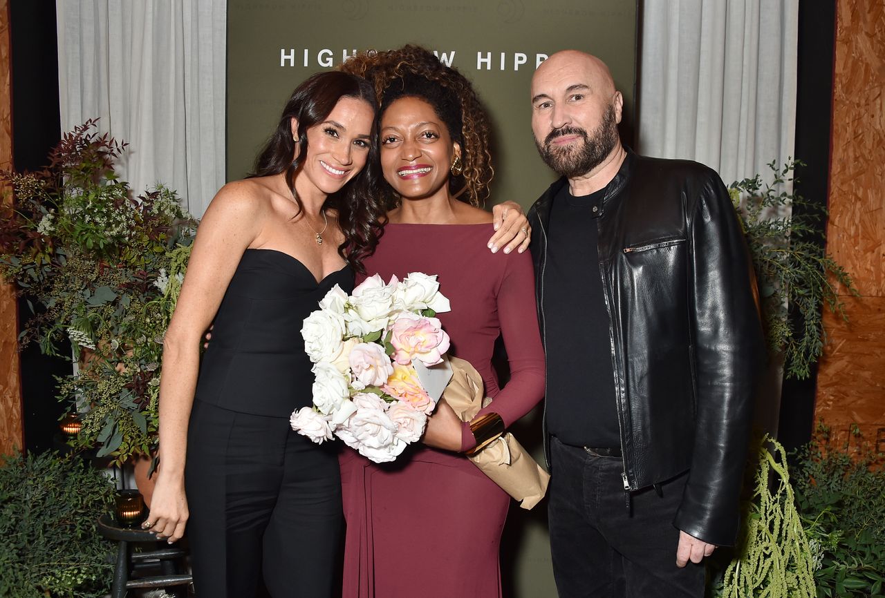 Meghan Markle wearing a black corset and pants with her arm around Kadi Lee who is holding a bouquet of roses, and Serge Normant standing next to them in a black jacket