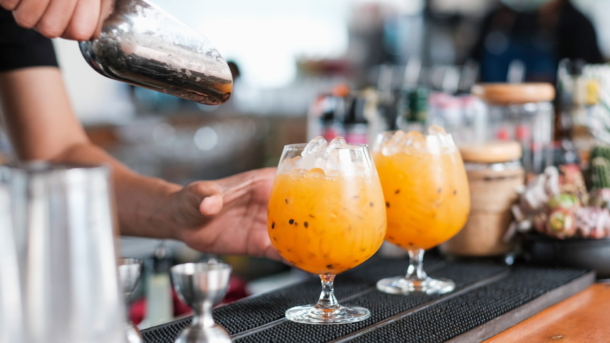side shot of a bartender pouring ice into two big bulbous glasses filled with a passionfruit cocktail