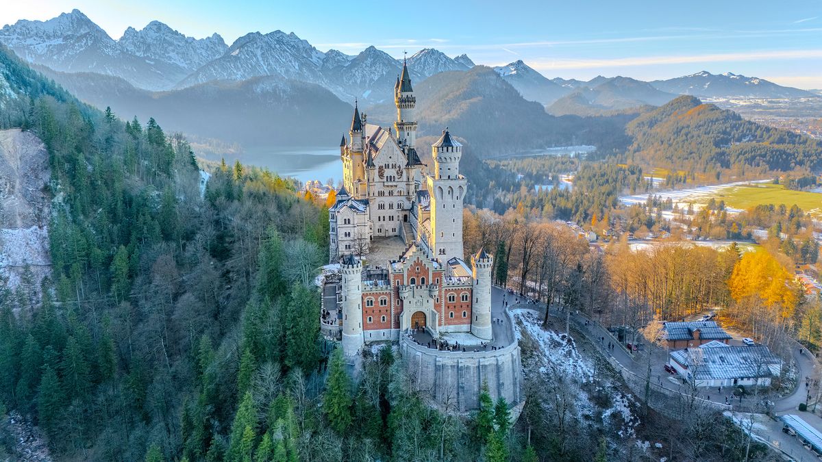 Aerial image of Neuschwanstein Castle during golden hour, with snow-capped mountains in background 