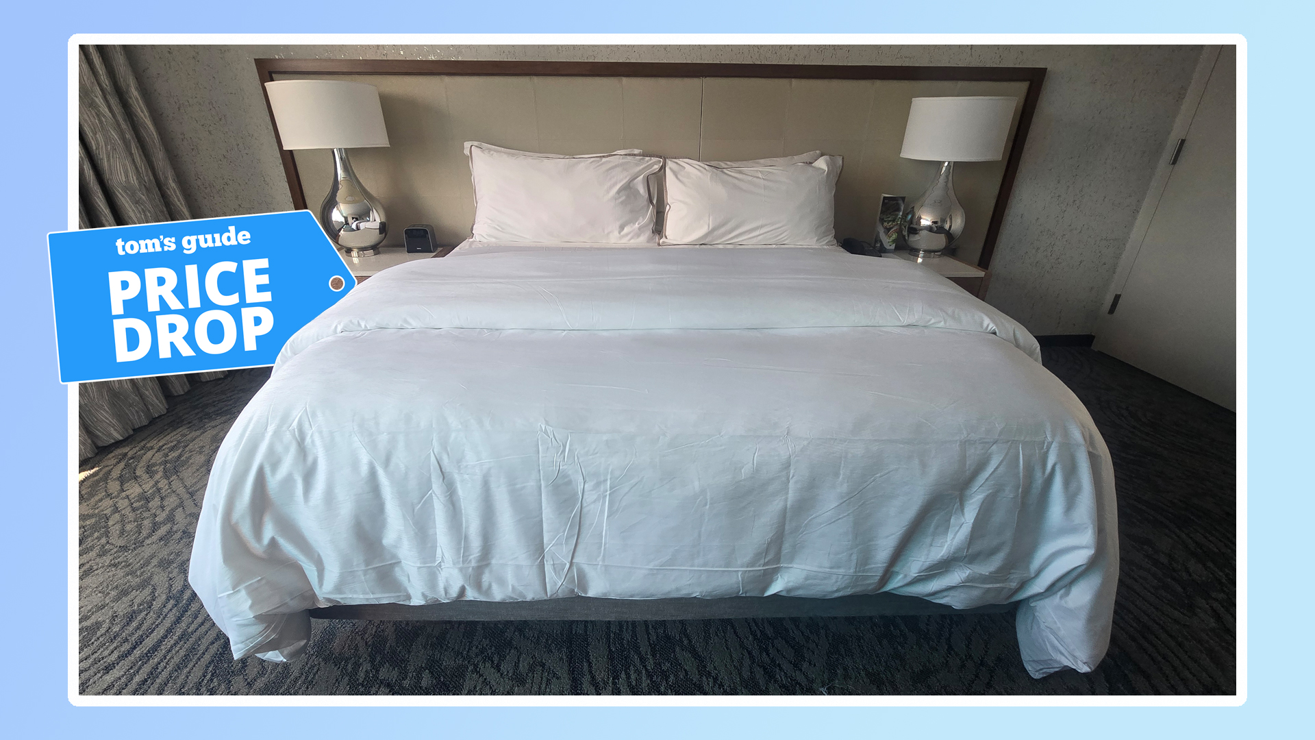 The Westin Heavenly Bed Mattress on a bed frame dressed in white bedding, in a Westin Hotel room. A Tom's Guide price drop graphic sits on the left-hand side and a blue border surrounds the picture