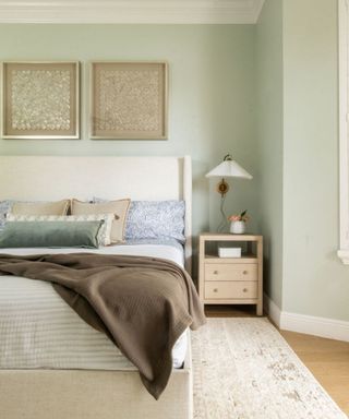 A mint green bedroom with a nightstand, bed with pillows and blankets on, and wall art