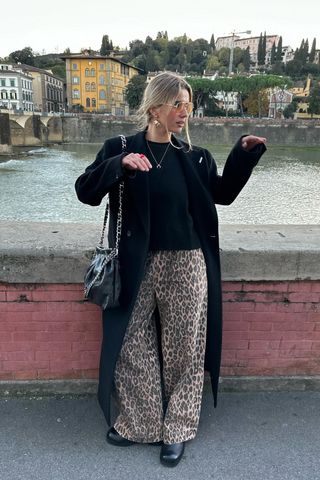 Eine Frau geht den Bürgersteig entlang und trägt einen langen schwarzen Mantel, einen schwarzen Pullover mit Rundhalsausschnitt, eine Hose mit Leopardenmuster und schwarze Stiefeletten.