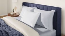 Three Tempur-Pedic cooling pillows shown on a dark blue fluted bed frame with white bedding and blue blankets, beside a wooden nightstand with a clock, candle, and two small vases. The room is white.
