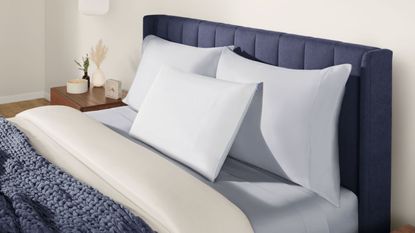 Three Tempur-Pedic cooling pillows shown on a dark blue fluted bed frame with white bedding and blue blankets, beside a wooden nightstand with a clock, candle, and two small vases. The room is white.