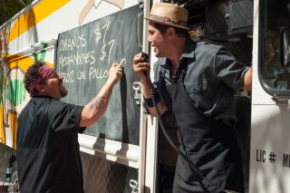 (L to R) Jon Favreau as Carl Casper writing a menu on the side of a food truck as John Leguizamo as Martin talks into a mic in Chef