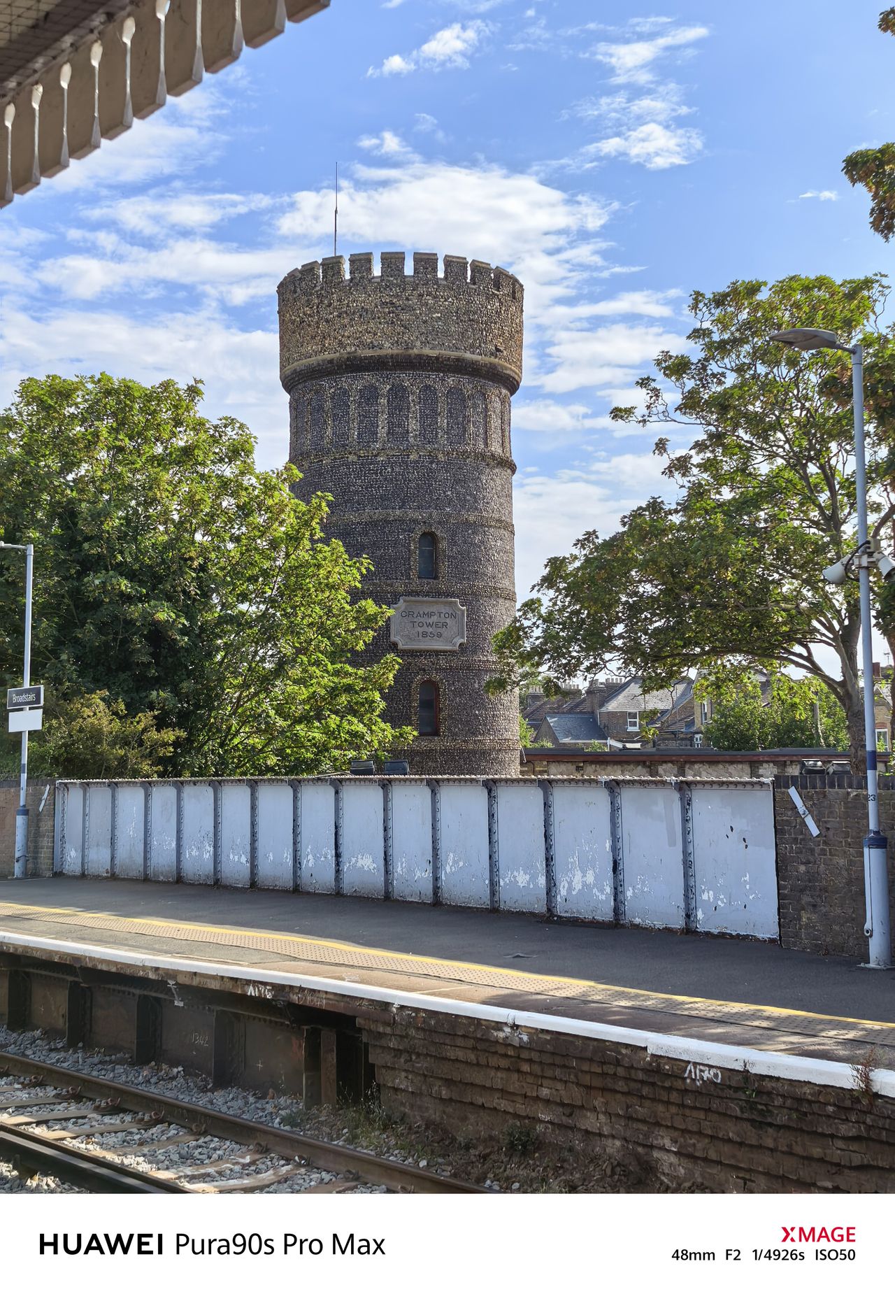Historic Crampton Tower beside a railway platform, framed by leafy trees under a blue sky.